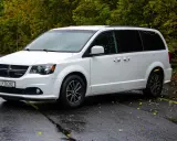 Biały Dodge Grand Caravan na mokrej drodze, otoczony jesienną zielenią.