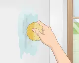 Mano limpiando una mancha en una pared blanca con una esponja. Aprende como limpiar paredes blancas fácilmente.