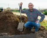 Un homme en chemise à carreaux et jean, pelle à la main, près d'un tas de tourbe blonde. Il est prêt pour son utilisation au jardin.