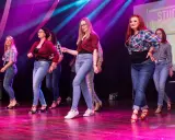 Grupa kobiet w kratę i jeansach tańczy na scenie. Występ latino solo dla początkujących.