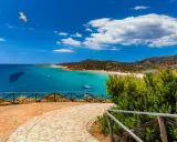 Vista panoramica di una baia con acque turchesi, spiagge affollate e montagne verdi. Un gabbiano vola nel cielo azzurro sopra Quartu e dintorni.