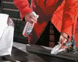 Osoba w pomarańczowej kurtce pokazuje, jak czyścić biżuterię ze stali chirurgicznej, używając sprayu i papierowego ręcznika.