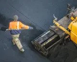 Arbeiter schaufelt heißes Bitumen auf eine Straße, während ein Asphaltfertiger das Material verteilt.