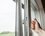 Mano girando manija de ventana blanca, asegurando la permeabilidad al aire clase 4. Ventana abierta con vista al exterior.