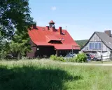 Chatka Górzystów z czerwonym dachem i drewnianymi budynkami. W tle las, a przed budynkami zielona łąka.
