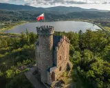 Kamienna wieża z polską flagą na tle jeziora i gór. Wspaniałe **cieplice atrakcje** dla miłośników historii i przyrody.