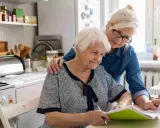Dos mujeres mayores revisan documentos en la cocina. Una apoya a la otra, quizás tramitando la pensión de orfandad mayores de 30 años.