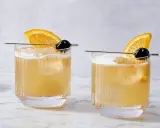 Dwa drinki Rusty Rat z pomarańczą i wiśnią na patyczku, podane w szklankach z lodem.