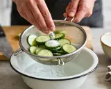 Préparation d'un concombre à la crème : des rondelles de concombre sont salées dans une passoire au-dessus d'un bol d'eau.