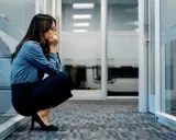 Frau kauert im Büroflur, Hände vor dem Mund, die Augen geschlossen. Sie scheint von **Angst vor der Arbeit** überwältigt zu sein.