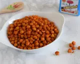 Un tazón blanco lleno de garbanzos fritos crujientes y especiados. Una cuchara de madera decorada y una lata de especias están al fondo.