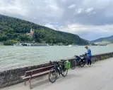 Mann packt Gepäck für eine Fahrradtour am Rhein. Im Hintergrund Burg und Fluss.