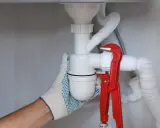 Ręka w rękawicy dokręca kluczem hydrauliczny syfon samoczyszczący, co znaczy, że zapobiega on zatorom.