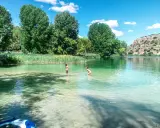 Dos niños juegan en las cristalinas aguas de las Lagunas de Ruidera, rodeados de vegetación y un paisaje montañoso.