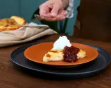 En bit krämig ostkaka med keso, toppad med vispgrädde och sylt. Serveras på en orange tallrik.