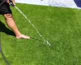 Persona riega césped artificial con manguera. Aprende cómo limpiar césped artificial fácilmente para mantenerlo verde y fresco.