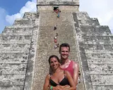 Coppia sorridente davanti alla piramide di Kukulkan, con turisti che salgono le scale del chichen itza interno.