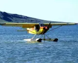 Gelbe Piper Super Cub mit Schwimmern auf ruhigem Wasser, im Hintergrund eine grüne Hügelkette.