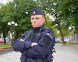 Policjant w granatowym mundurze, w sprawie umundurowania policjantów, stoi na tle zieleni.