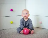 7 miesięczne dziecko z niebieskimi oczami siedzi na dywanie, trzymając w rączkach różową piłeczkę.