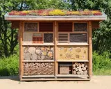 Un hotel de insectos con techo verde, lleno de materiales naturales como troncos, cañas y ladrillos, ofrece refugio a pequeños animales.