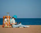 Dziewczyna w niebieskim kapeluszu siedzi na plaży z walizką, laptopem i telefonem, planując, jak tanio podróżować.