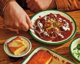 En hand tar en sked ur en skål med mustig texas chili, toppad med jalapeños och gräddfil. Bredvid ligger gyllene majsbröd.