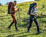 Trzy osoby wędrują po zielonym zboczu. Mężczyzna i kobieta w wygodnych, sportowych spodniach w góry, z plecakami i kijkami trekkingowymi.