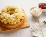 Rosca de Pascua decorada con crema pastelera y azúcar granulada, lista para disfrutar. A su lado, cuencos con cerezas y ralladura, y un batidor.