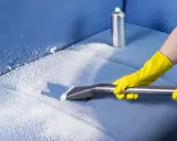 Mani con guanti gialli usano un aspiratore per pulire un divano in microfibra con schiuma detergente.