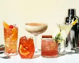Eine Auswahl an fruchtigen Cocktails, garniert mit Orangen, Äpfeln und Kirschen, neben einem Cocktail-Shaker.