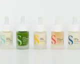 Cinq flacons de sérum et crème hydratante aux couleurs vives, chacun avec une étiquette indiquant son usage : anti-soif, imperfections, unifiant, anti-grisaille, anti-âge.