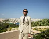 A man in a light tan suit, tie, and sunglasses offers inspiration for what to wear to a communion as a guest male.