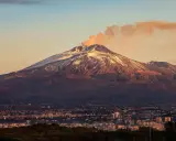 Etna fumante, con la cima innevata che brilla al tramonto, domina una città siciliana.