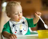 Małe dziecko z marchewkowym puree na buzi i śliniaczku próbuje samodzielnie jeść. Oto jak sprać marchewkę z ubranka!