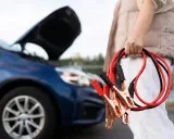 Osoba trzyma kable rozruchowe. Z przodu niebieski samochód z otwartą maską, sugerujący problem z akumulatorem.