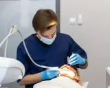 Dentysta w niebieskim fartuchu i masce wykonuje zabieg. Pacjentka ma założone okulary ochronne. Nowoczesne znieczulenie komputerowe zapewnia komfort.