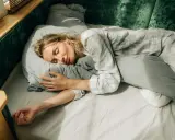 En kvinna sover lugnt i sängen, omfamnande en kudde. Solstrålar faller in genom fönstret och skapar en känsla av guidad avslappning.
