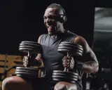Uomo con cuffie nella palestra gym, solleva pesi con musica per allenamento. Grinta e determinazione.