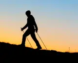 Sylwetka osoby uprawiającej nordic walking na tle zachodzącego słońca. Czy wolisz nordic walking rano czy wieczorem?
