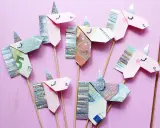 Kreatives Geld falten: Mehrere Einhorn-Figuren aus Geldscheinen und Glitzerpapier auf Holzstäbchen vor rosafarbenem Hintergrund.