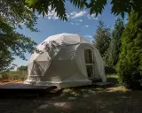 Biały namiot kopułowy, idealny na glamping noclegi, otoczony zielenią. Drewniany taras z meblami zachęca do relaksu.