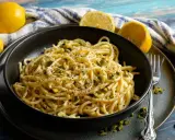 Eine Schüssel mit Zitronen Parmesan Pasta, garniert mit gehackten Pistazien und schwarzem Pfeffer. Frische Zitronen liegen daneben.