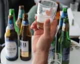 Ręka trzyma kieliszek z piwem bezalkoholowym. W tle kilka butelek piwa, idealne na przerwę w pracy.