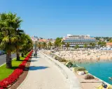 Sonniger Strandtag in Cascais bei Lissabon. Palmen säumen die Promenade, Menschen genießen das Meer und das Hotel Baía ragt empor.