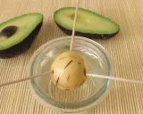 Préparation pour faire pousser un avocat : un noyau est suspendu dans un verre d'eau avec des cure-dents, à côté de deux moitiés d'avocat.