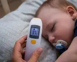 Elektroniczny termometr pokazuje 37.7°C, mierząc temperaturę niemowlaka śpiącego ze smoczkiem.