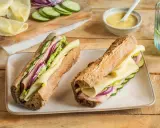 Deux sandwichs jambon fromage appétissants, garnis de concombre, oignon rouge et laitue, sur un plateau blanc.