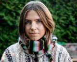 Elin Berlin med blå ögon och fräknar, klädd i en stickad tröja och randig halsduk, står framför en grön häck.