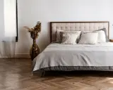 Cama con base tapizada o canapé, cojines mullidos y mantas grises. Un jarrón con pampas decora el rincón.
